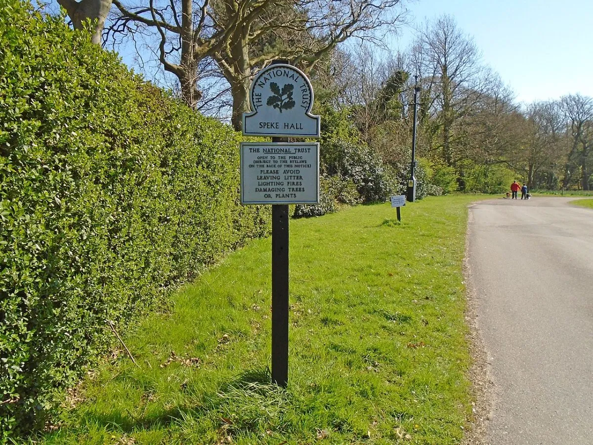 Een bezoeker die bij de ingang van een landhuis een pas of kaartje laat zien, of het bordje van English Heritage of het National Trust bij een toegangspoort.