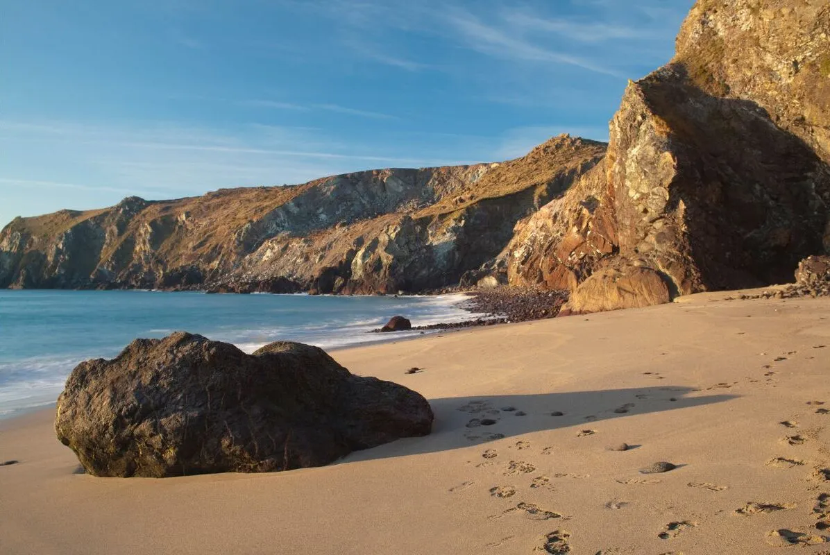 Kynance Cove bij hoog water, waar het zand volledig verdwenen is en de zee tegen de rotsen staat. Naast de herofoto bij laag water maakt dat het verschil meteen zichtbaar.